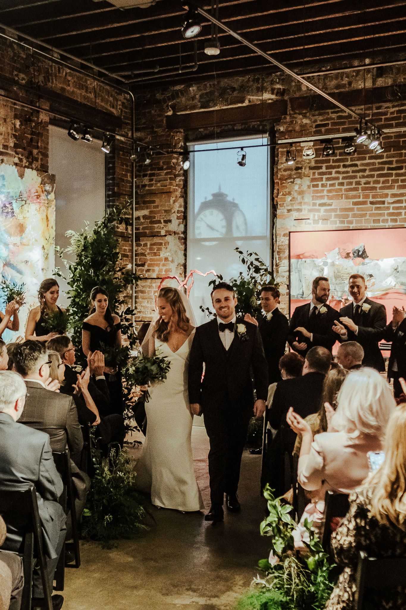 Wedding party exits down the aisle in an industrial venue with exposed brick walls and hanging greenery.