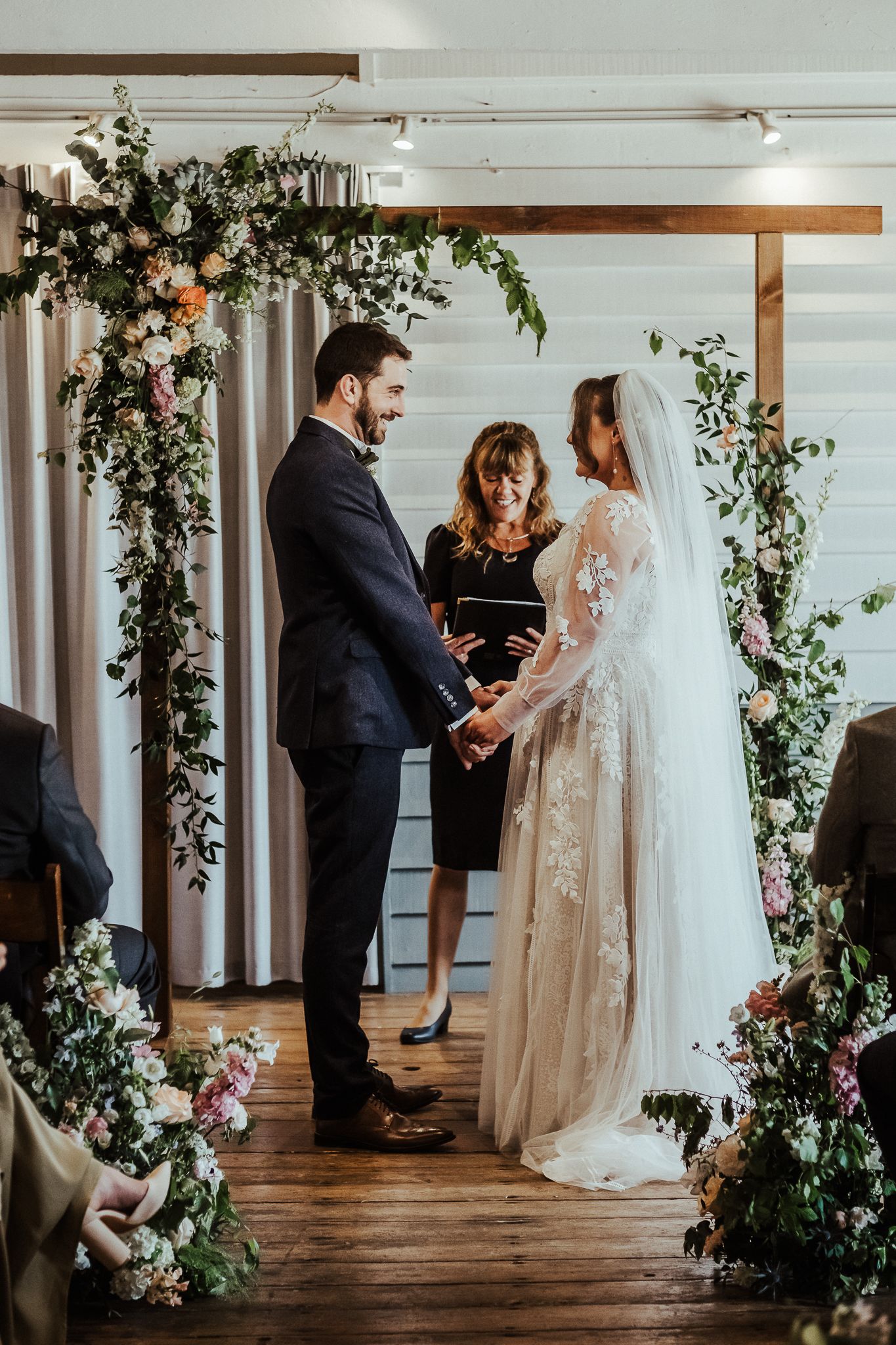 A couple stands beneath a floral arch during their indoor wedding ceremony with greenery and white flowers.