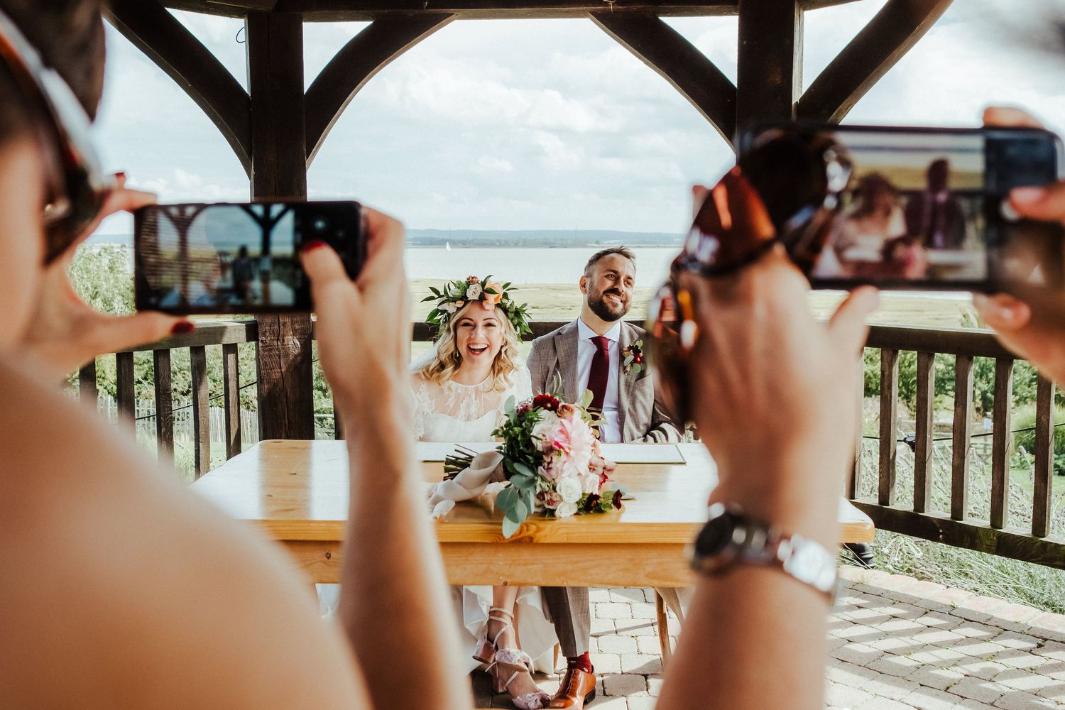 Wedding guests capturing photos under rustic wooden pavilion by the water.