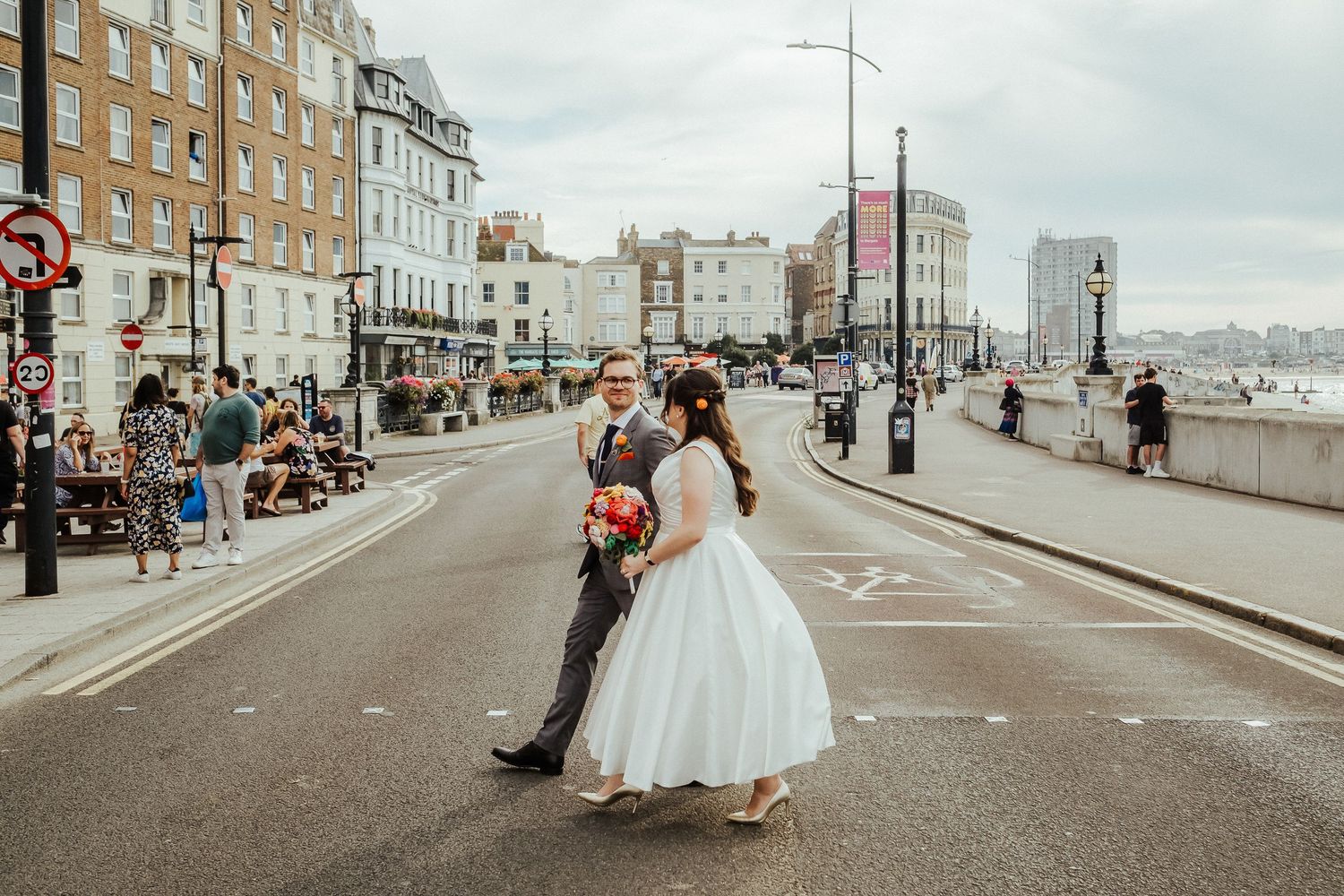 Newlyweds walking along seaside promenade with historic buildings.