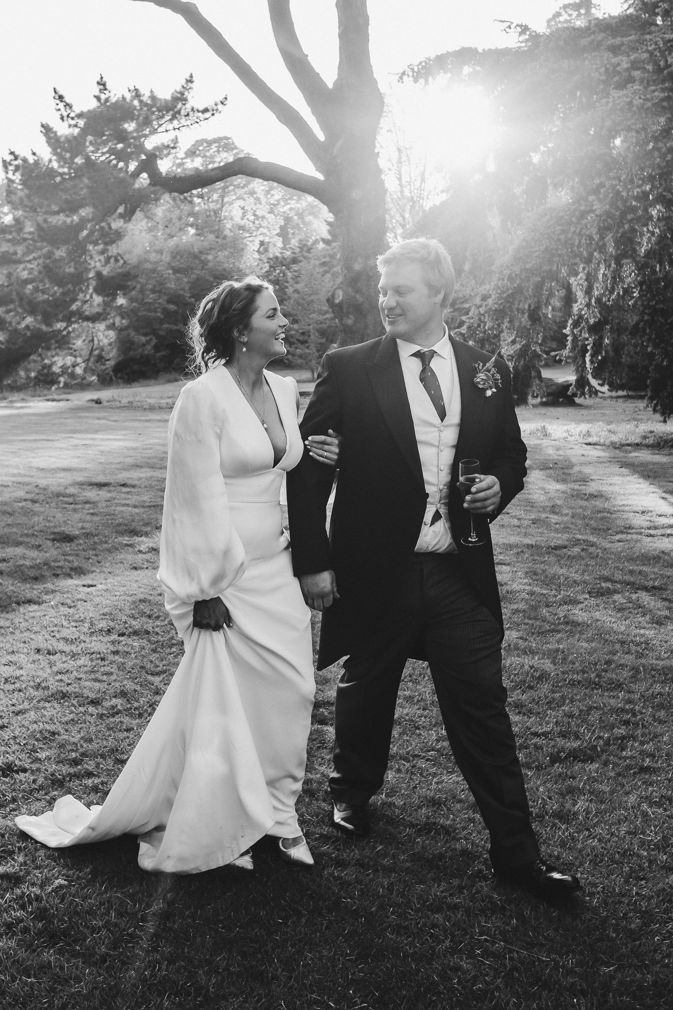 Couple walks across sunlit lawn during wedding celebration with sun flare through trees.