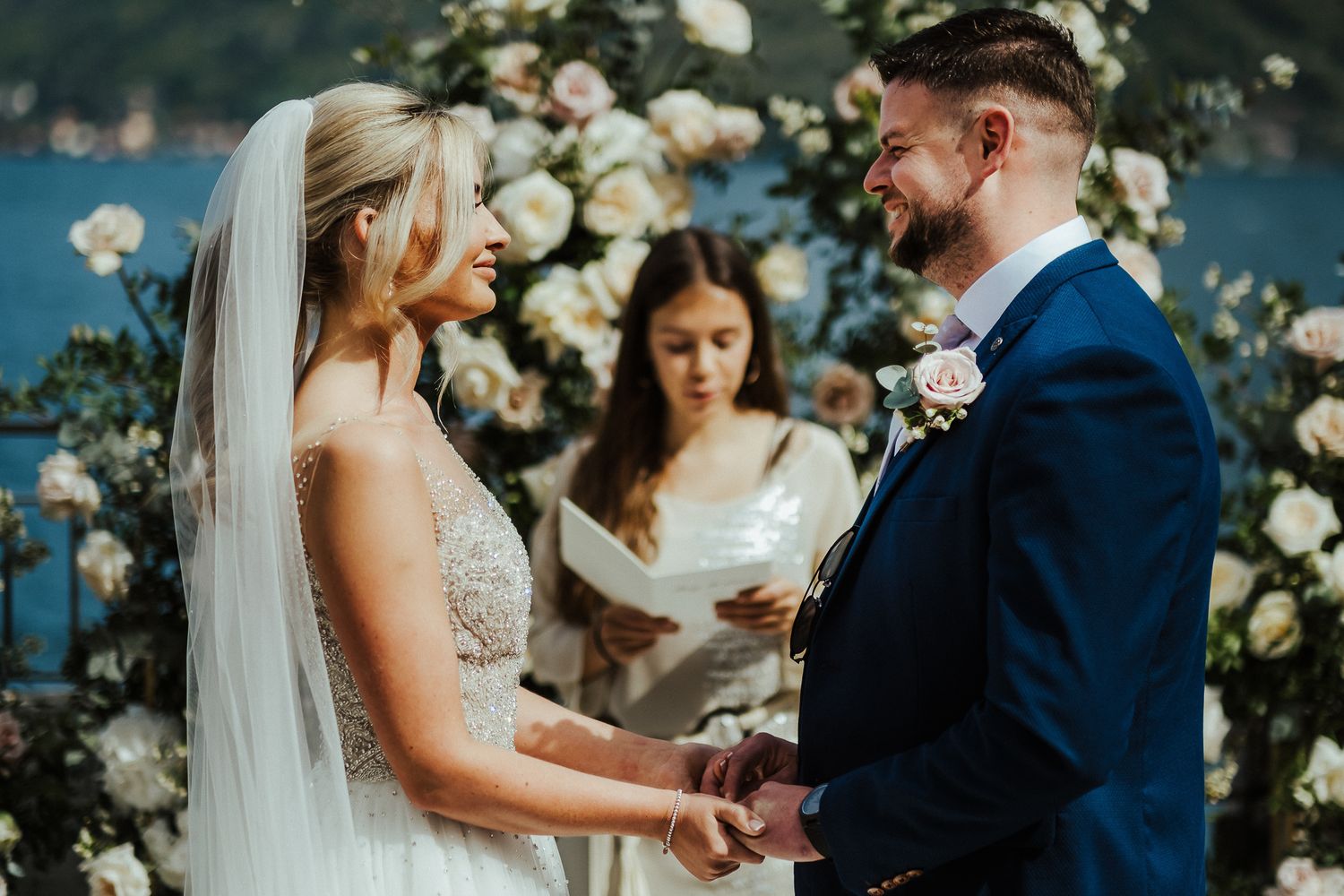 A lakeside wedding ceremony with a couple exchanging vows under a floral arch.