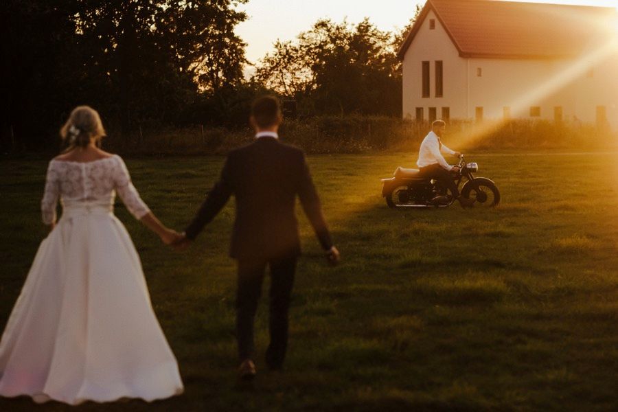 Newlyweds hold hands walking at sunset with a motorcycle and church in the background.