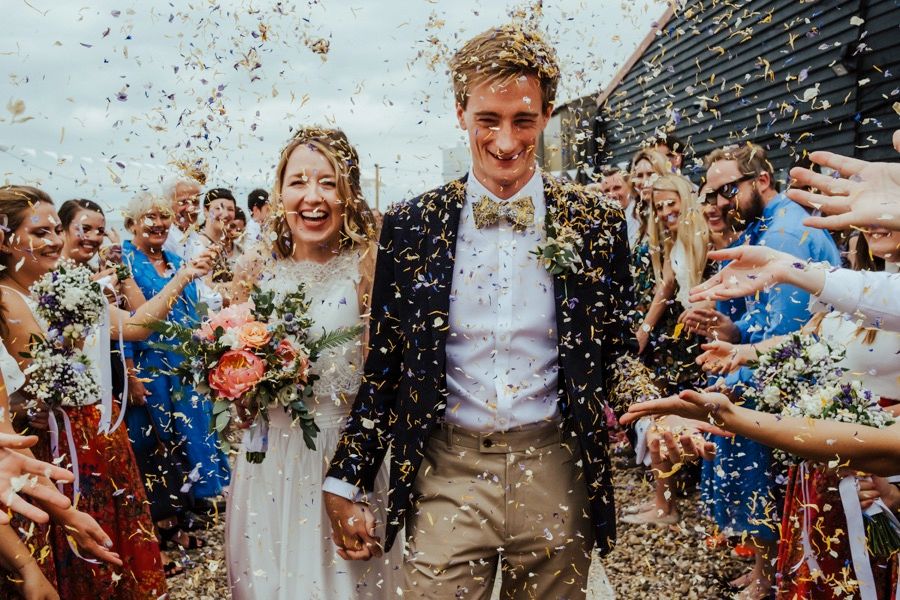 Newlyweds celebrate while guests throw colorful confetti during an outdoor wedding celebration.
