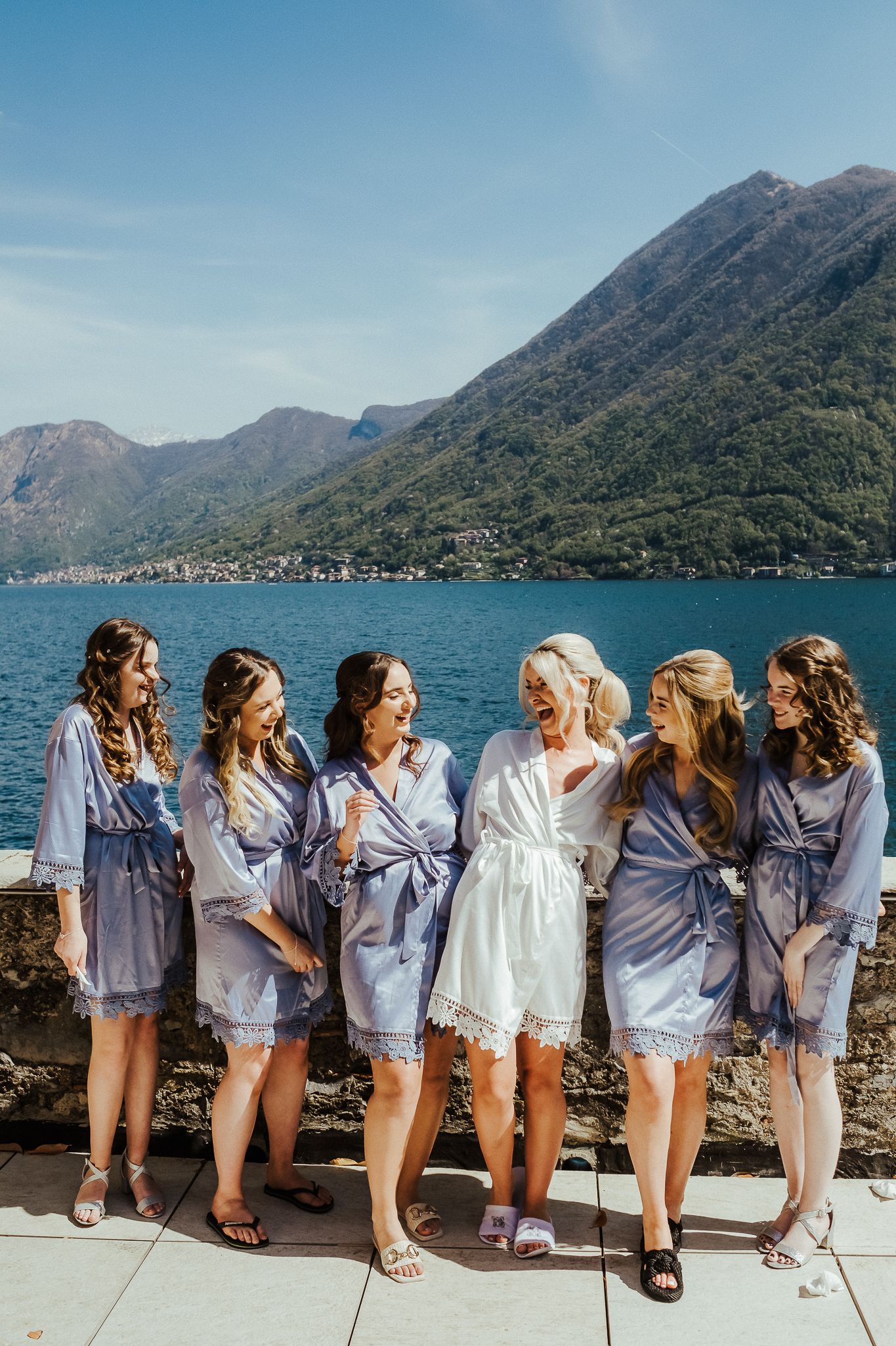 Bride and bridesmaids in matching robes pose together against stunning mountain lake backdrop.