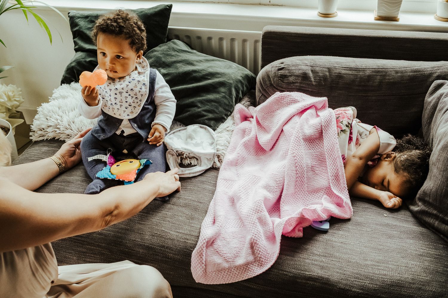 Toddler plays on couch while someone naps under pink blanket.