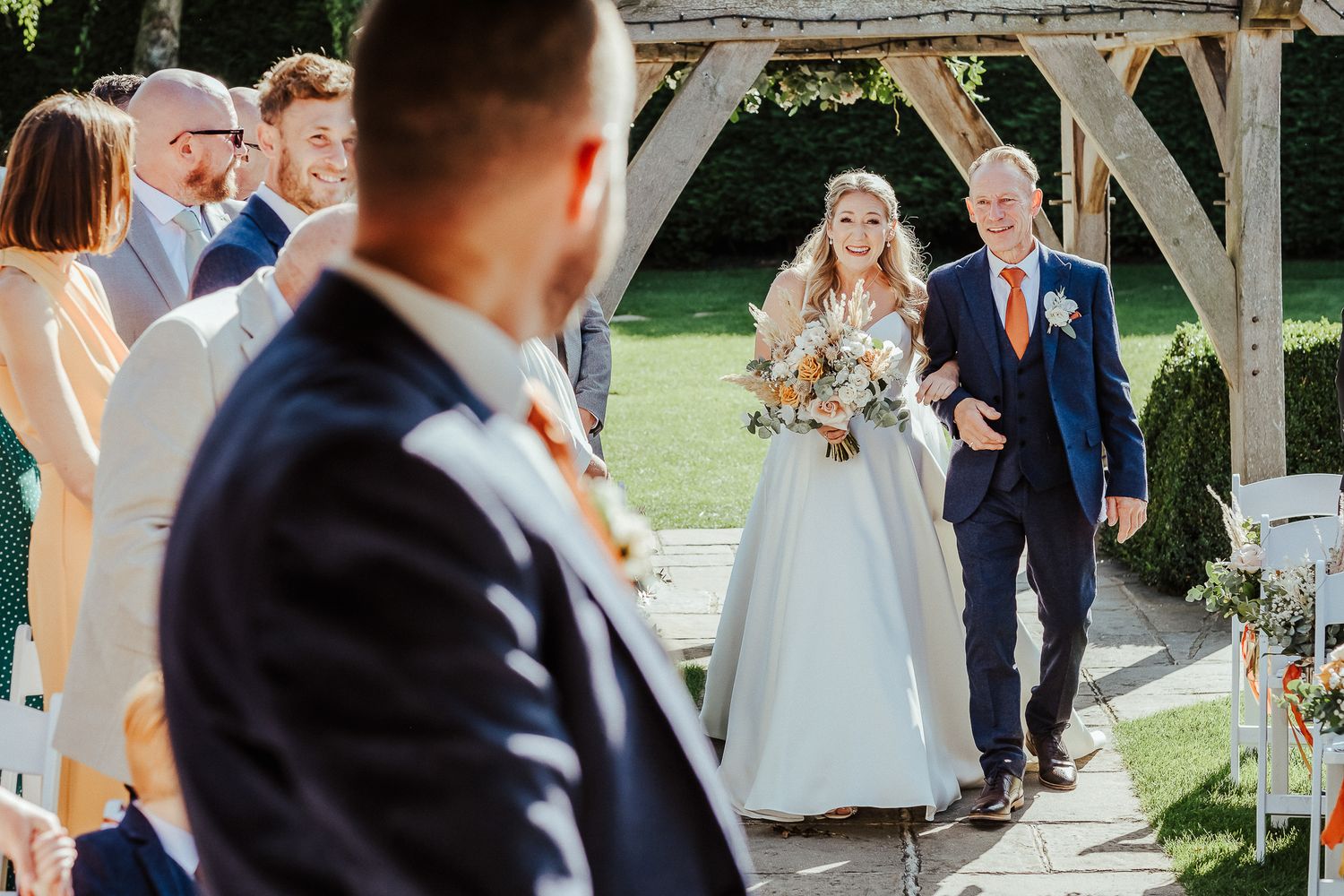 Bride walks down outdoor aisle with companion under rustic wooden gazebo.