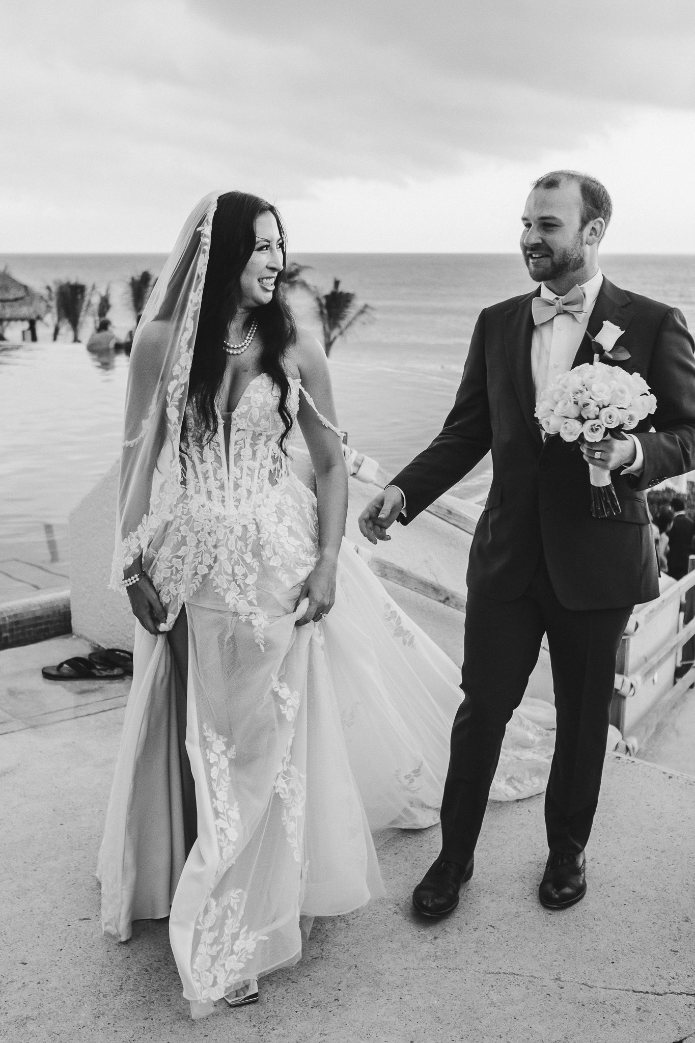 Joyful wedding couple sharing a candid moment while walking along an oceanfront path in black and white.