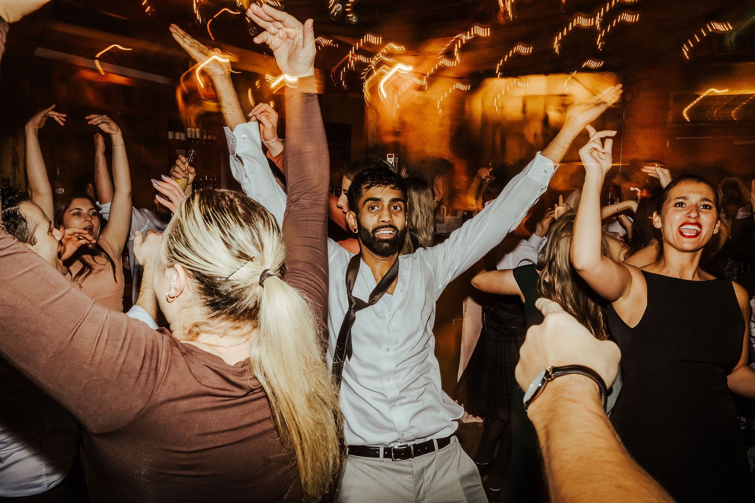 Energetic party crowd dancing with raised arms at a dimly lit reception venue with warm lighting and motion blur.