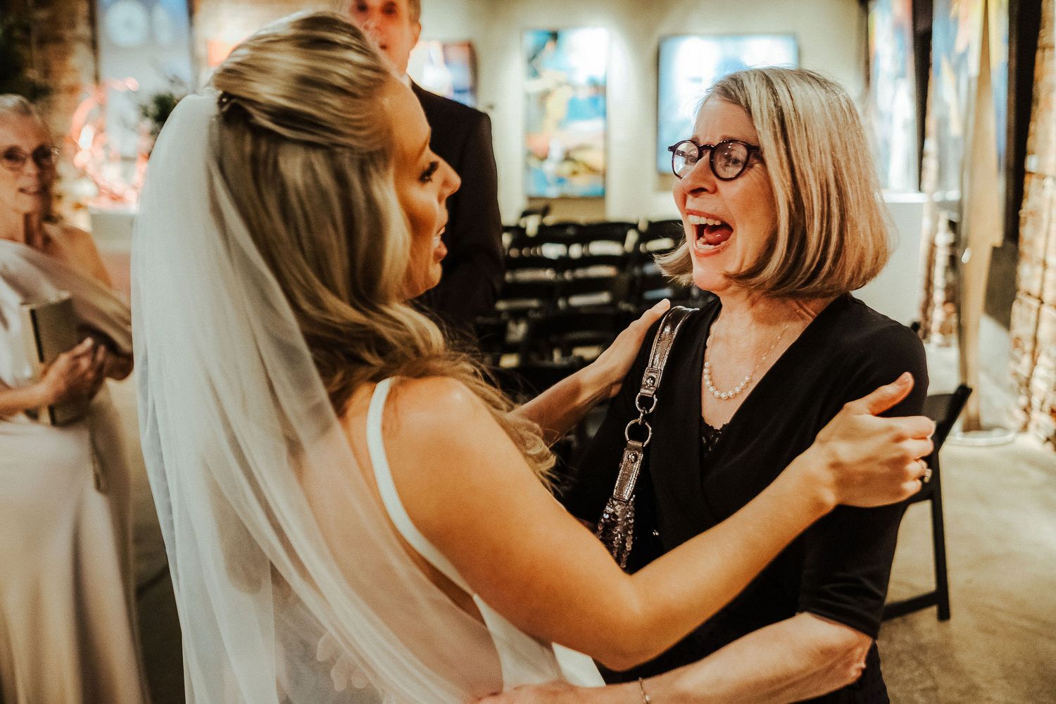 Bride in white veil sharing excited moment with wedding guest at indoor reception.