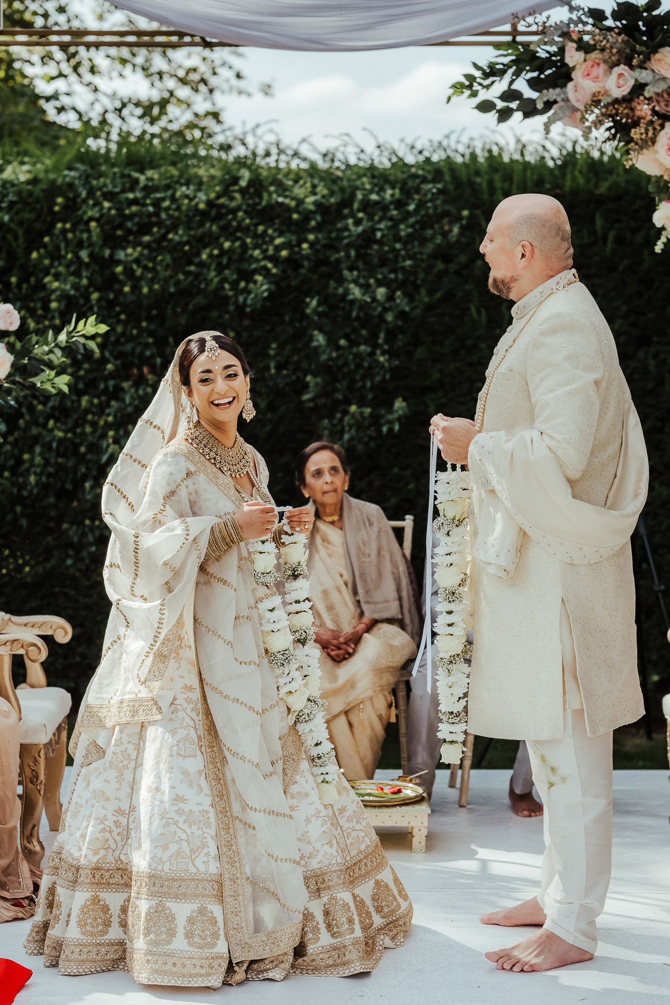 A couple in traditional white and cream Indian wedding attire performs a ceremony on an outdoor stage with floral decor.