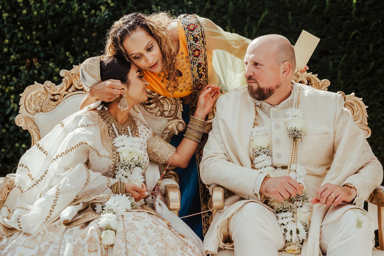 Three people in ornate white and gold royal attire share an intimate moment on an elegant baroque style settee.