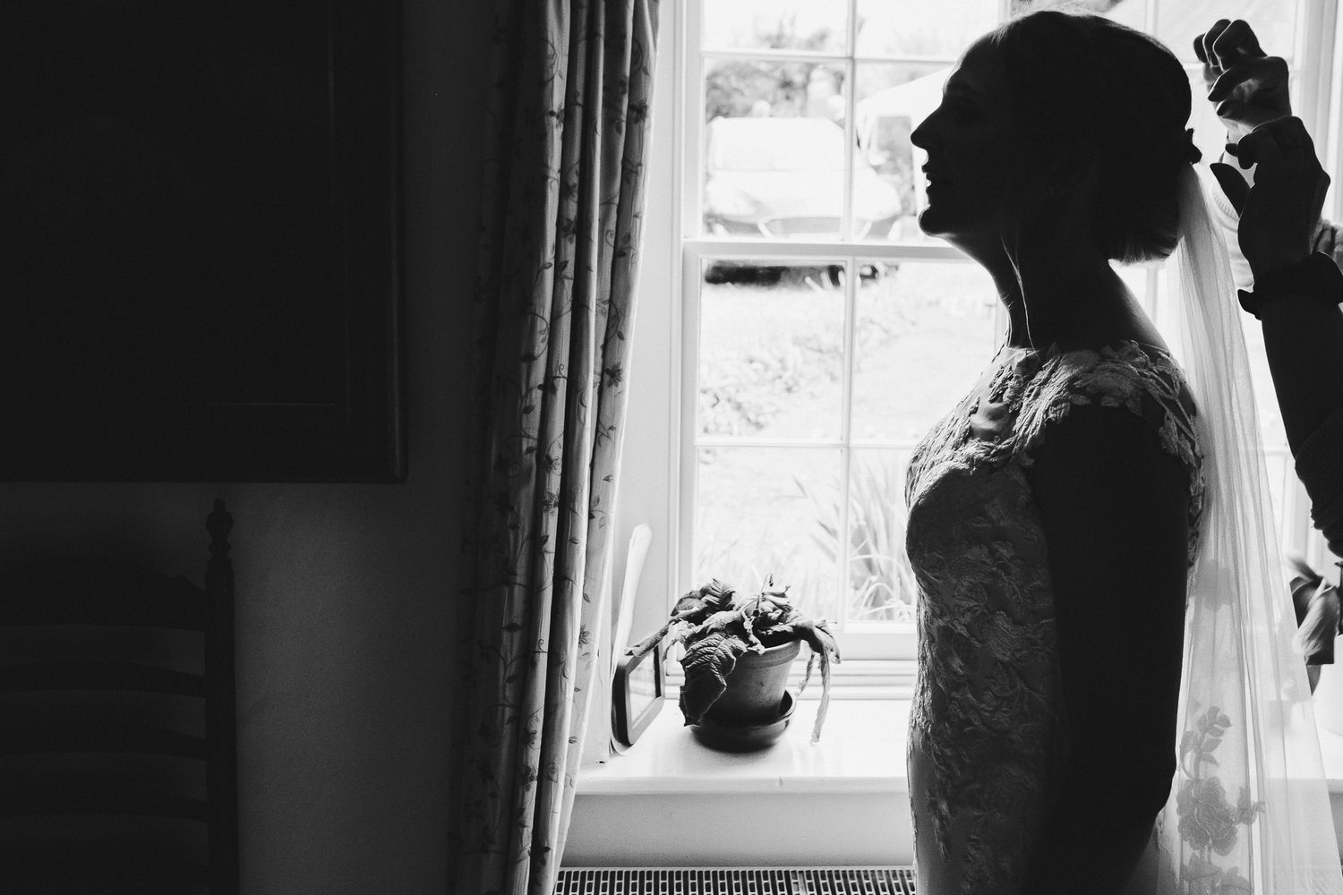 Silhouette of a bride getting ready by a window in a lace dress on her wedding morning.