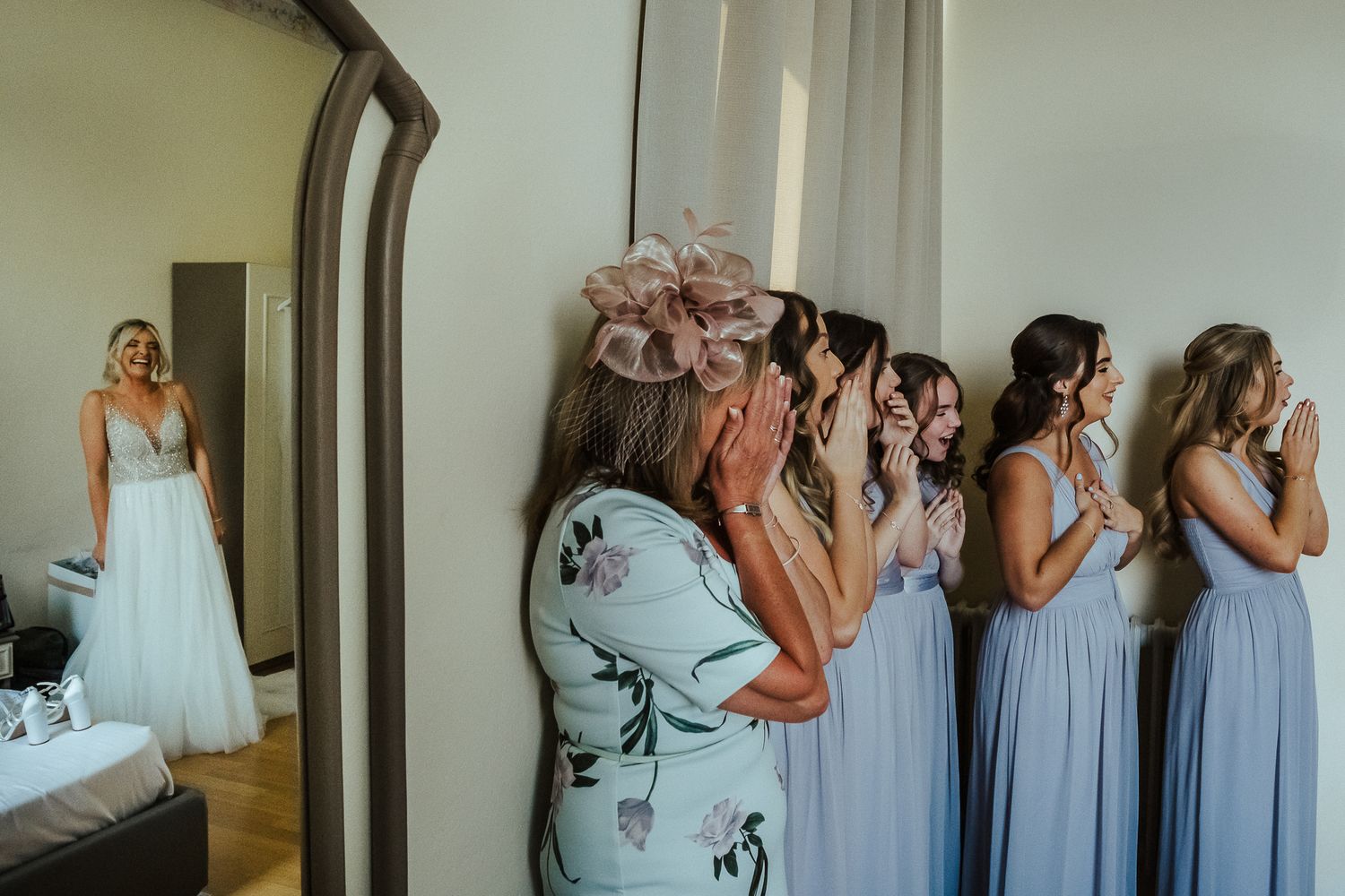 Emotional bridesmaids react as bride reveals her wedding dress in getting ready room.