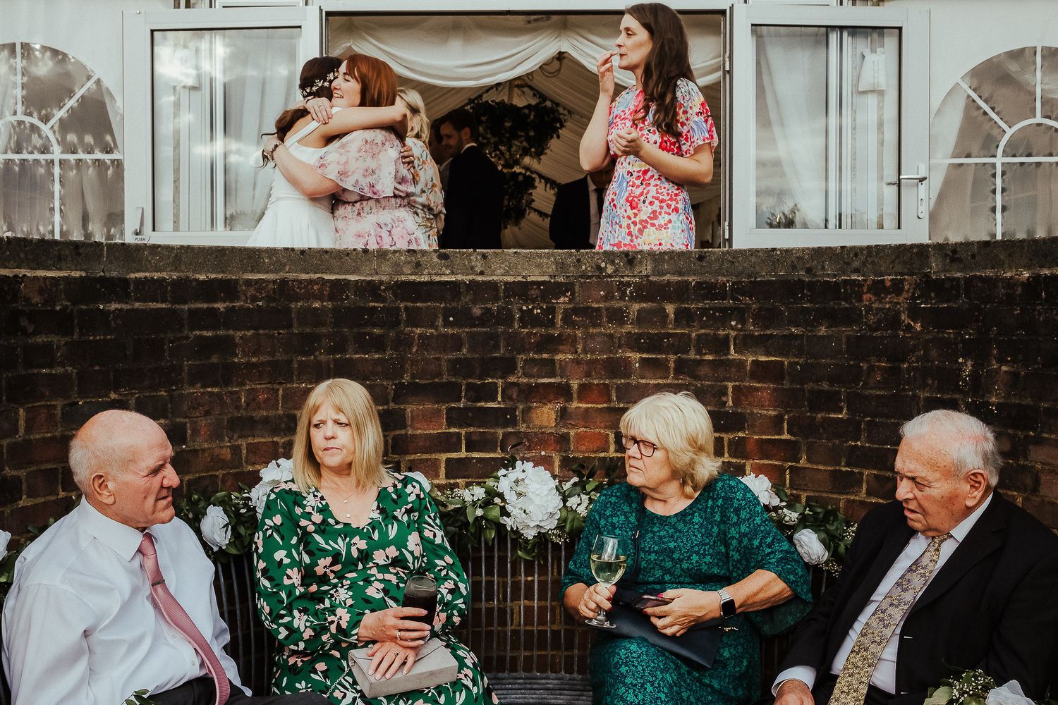 Guests enjoying an outdoor party with two people chatting above on a brick wall.