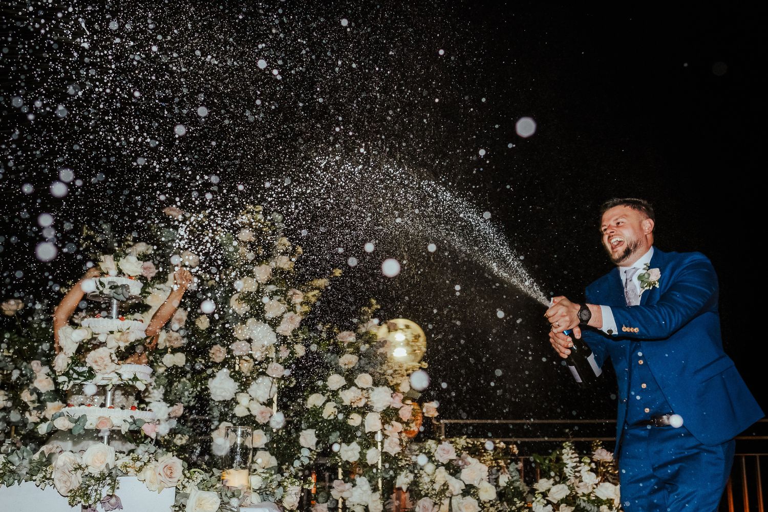 Champagne sprays dramatically against a dark night sky during wedding celebrations.
