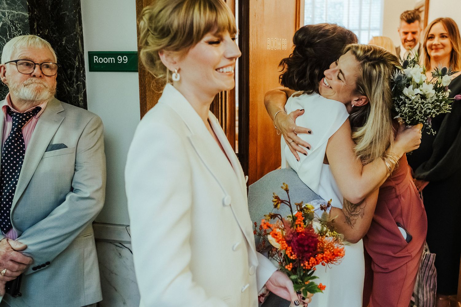 Joyful wedding gathering in hallway with celebratory hugs and bright floral bouquet.