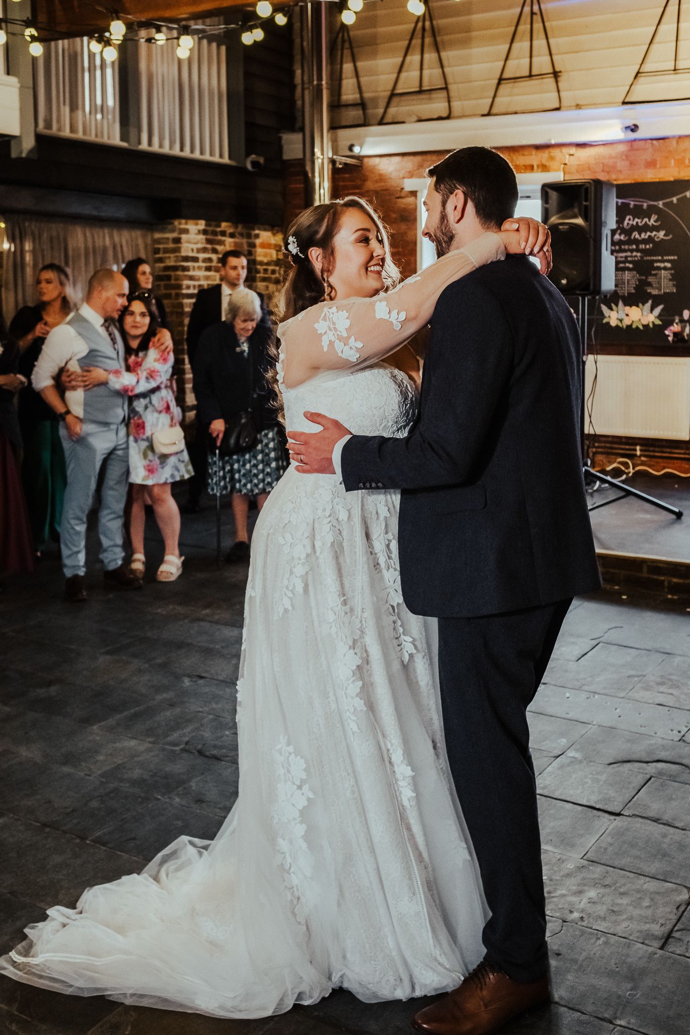 Newlyweds dance intimately in a rustic brick venue, with wedding guests watching in the background.