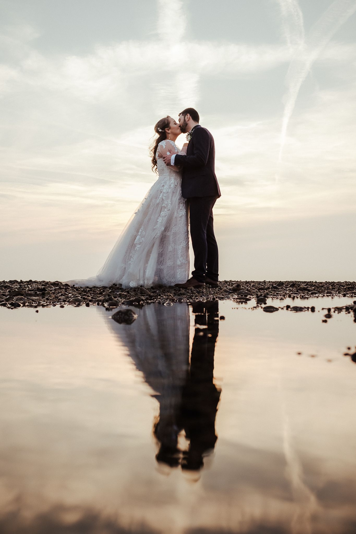 Romantic wedding couple embracing on a beach at sunset with their reflection in calm waters.