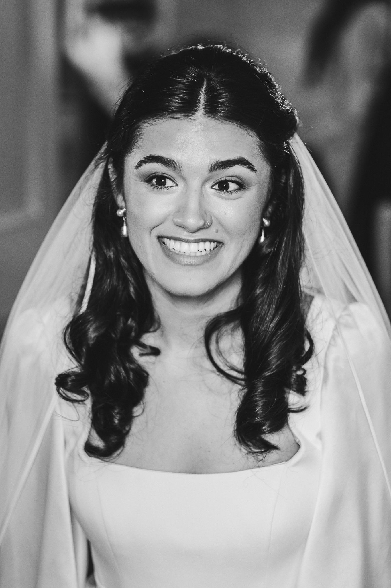 A radiant bride in a classic white dress with long dark curls and veil smiles at the camera.