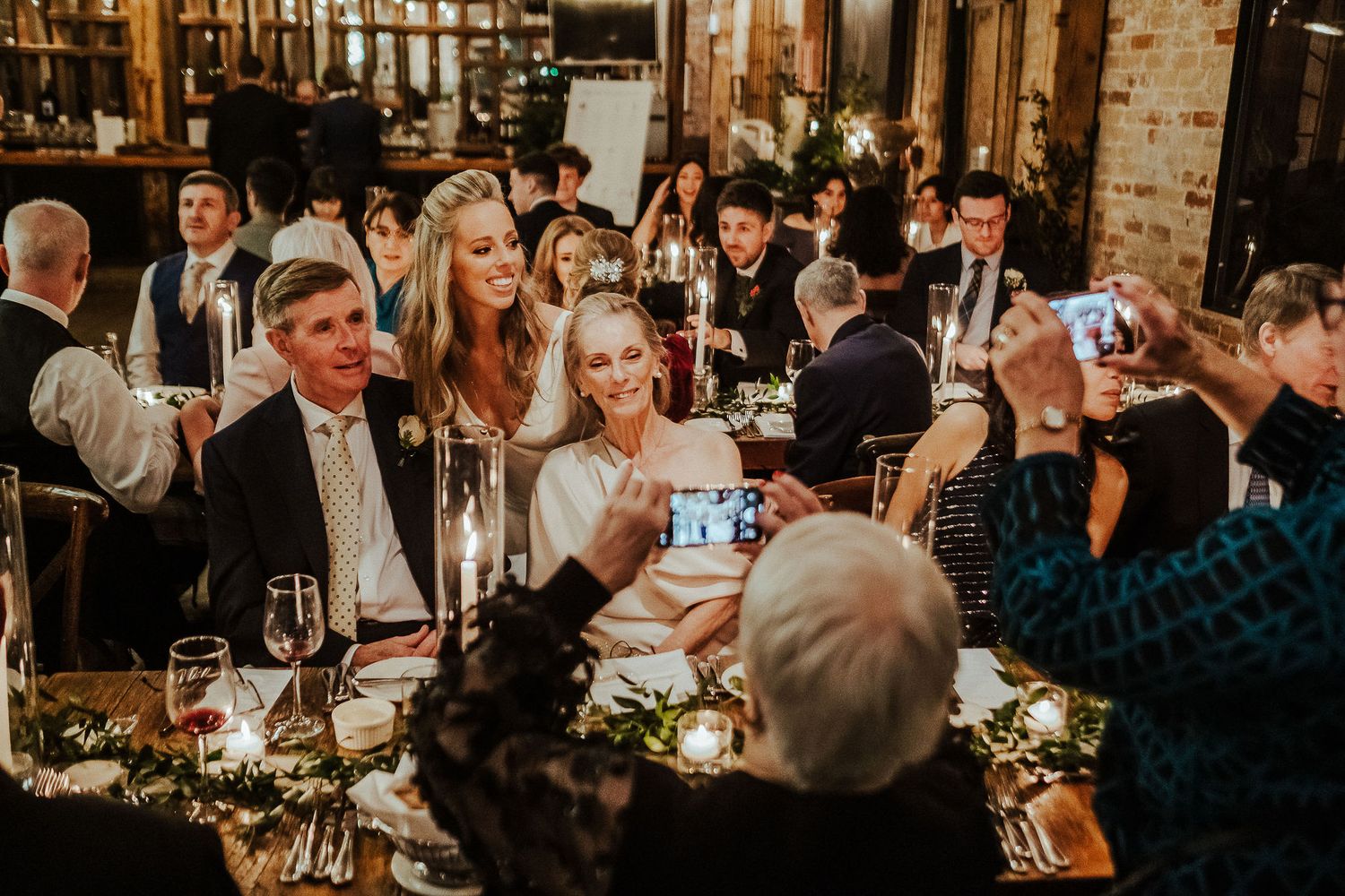 Warm evening reception with guests dining at decorated tables in rustic venue.