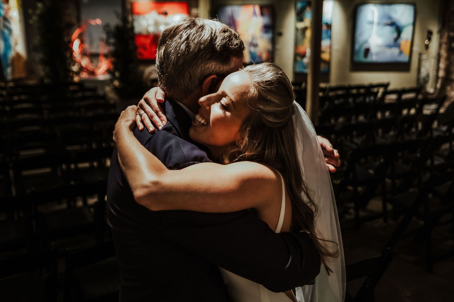 Intimate embrace between couple in dimly lit wedding venue with rows of chairs.
