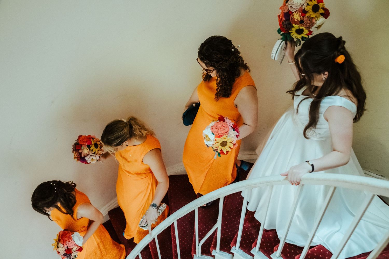 Bridesmaids in orange dresses and bride in white descend a spiral staircase holding bouquets.