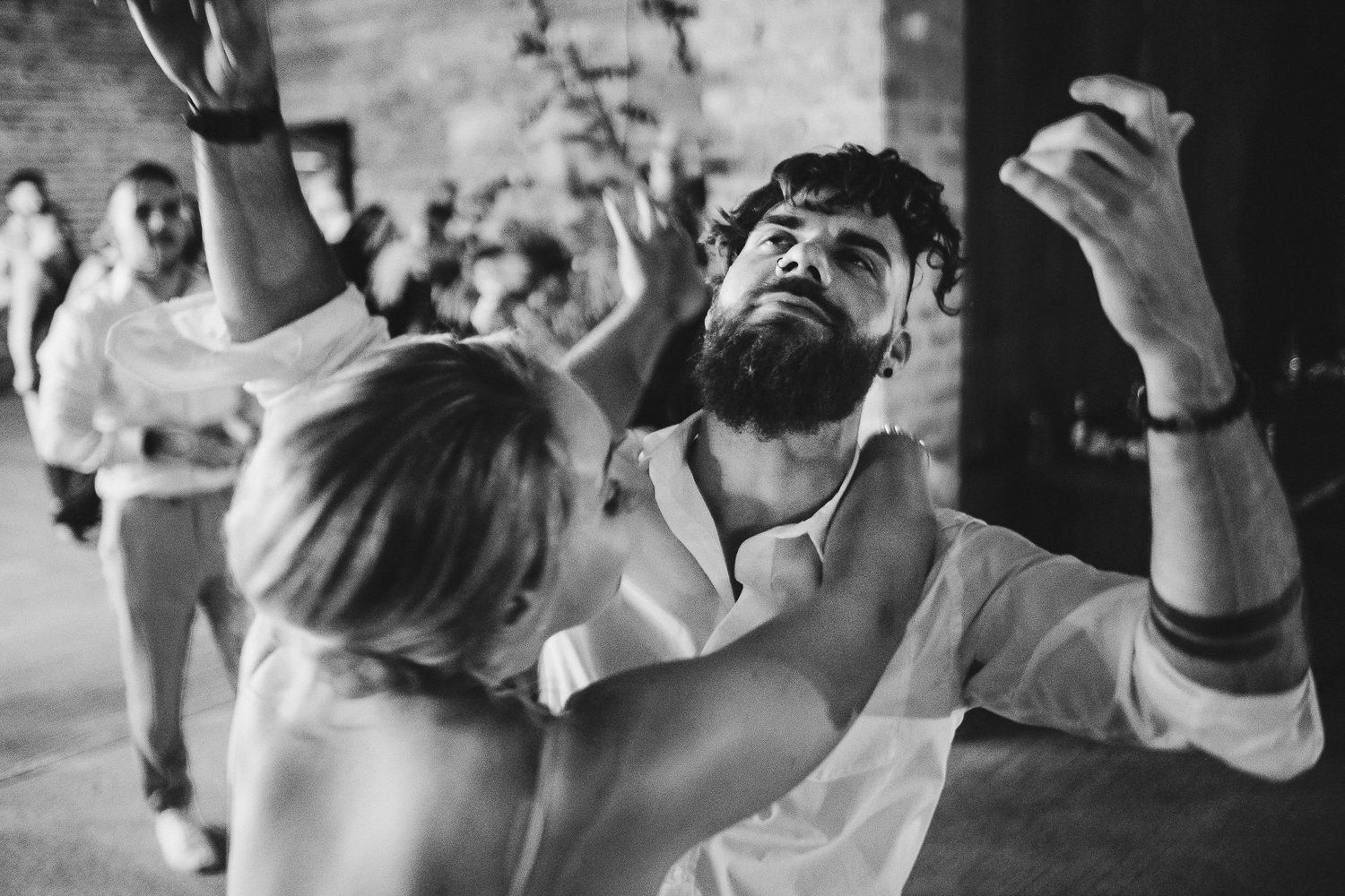 Candid black and white shot of wedding guests dancing with passionate expressions at reception.