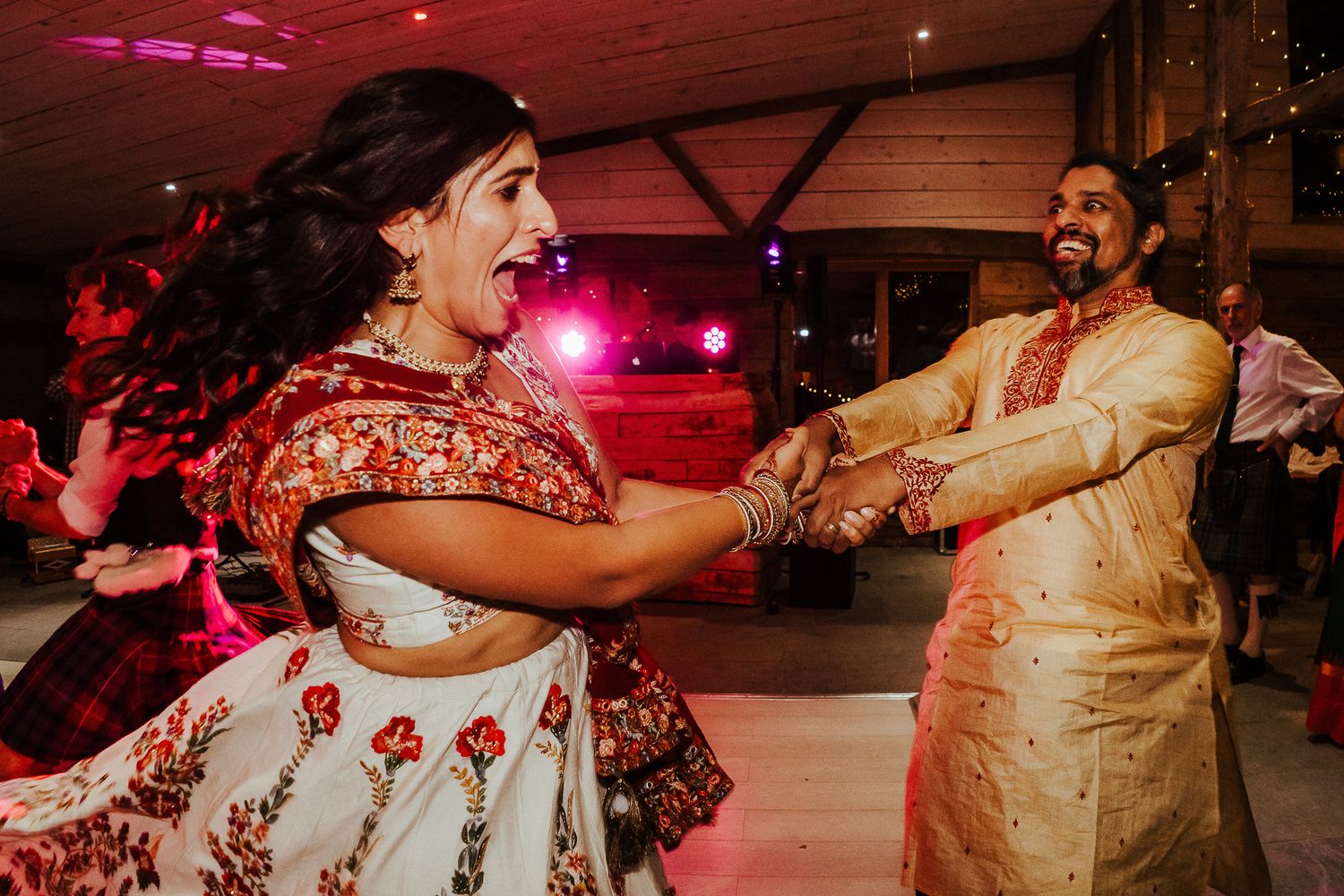 A couple dances joyfully at their Indian fusion wedding reception under red lighting.