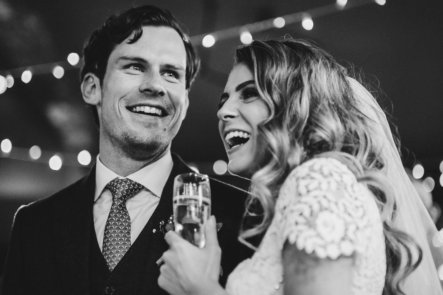 Black and white candid shot of couple sharing champagne and laughter during wedding celebration.