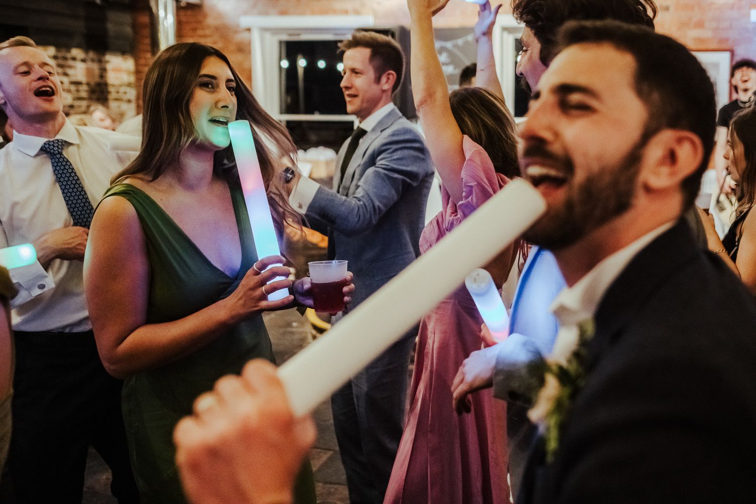Party guests wave colorful LED foam sticks while dancing at a nighttime celebration.