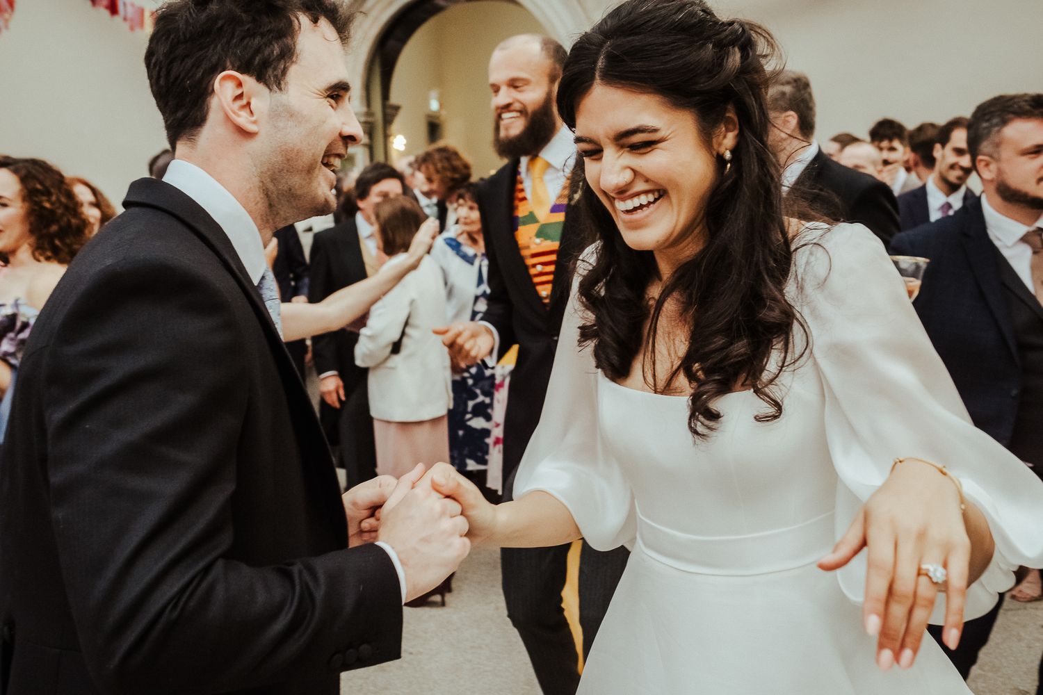 Couple dancing and laughing together during wedding celebration with guests in background.