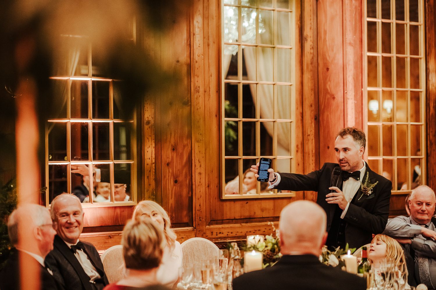 Wedding guests seated at wooden-paneled reception venue during evening speeches and toasts.