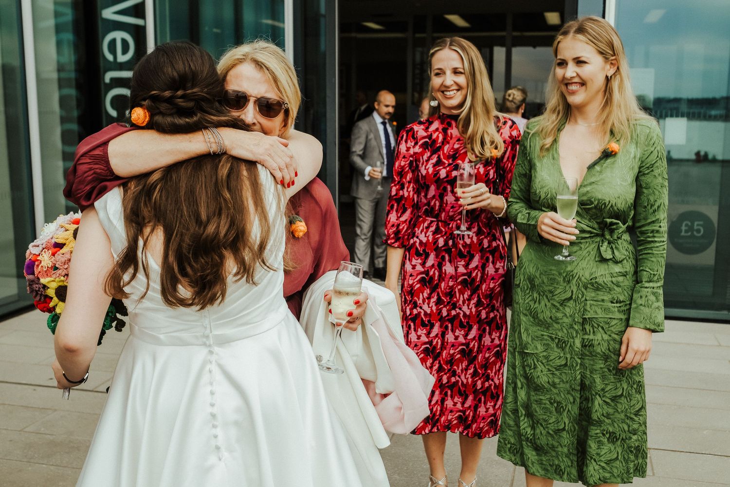 Wedding guests in colorful dresses greet and embrace the bride outside a modern building.