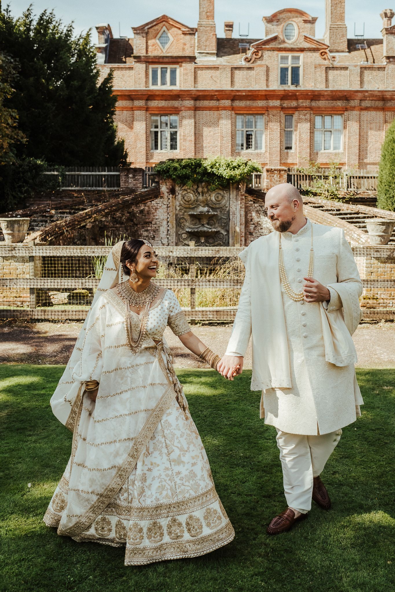 Traditional Indian wedding couple in cream-colored attire walking together on mansion grounds during outdoor celebration.