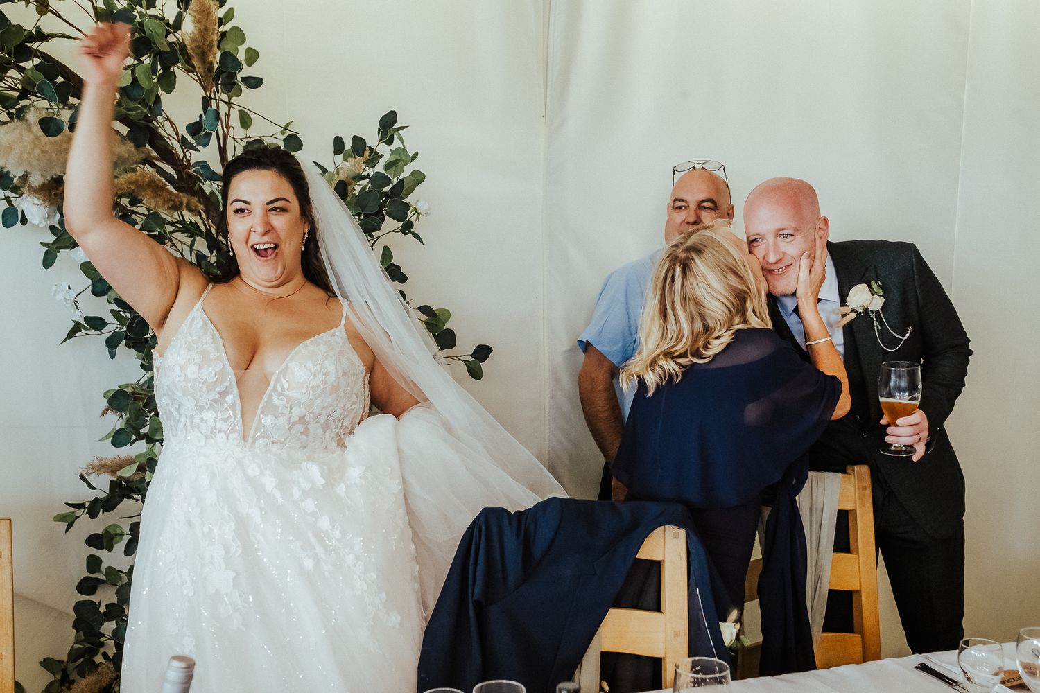 A bride in a lace wedding dress celebrates joyfully while guests enjoy drinks at a reception.