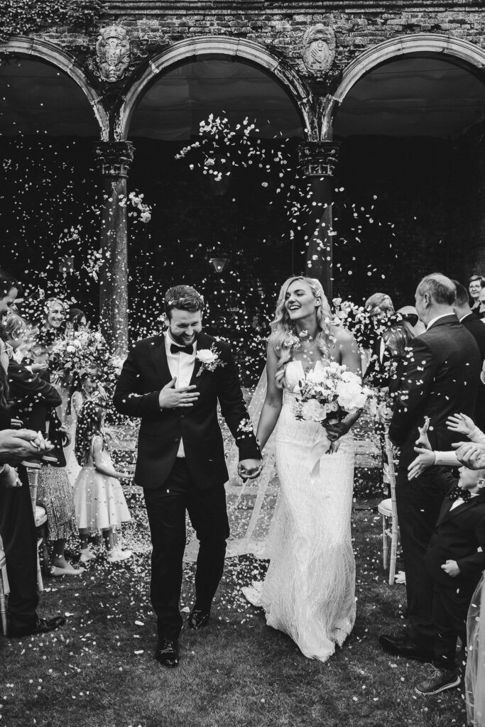 A newlywed couple walks under stone arches as guests shower them with confetti.