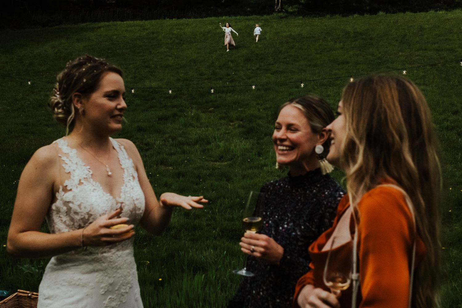 Three friends share laughter and drinks at an outdoor wedding reception with sheep grazing in the background.