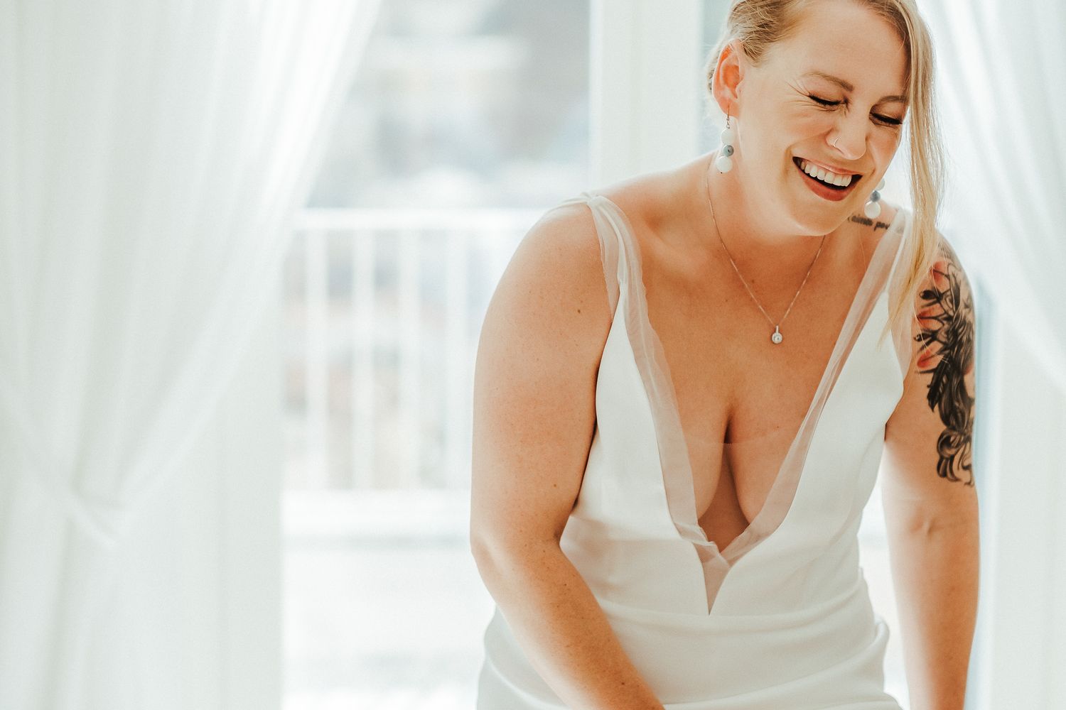 A person in a white wedding dress smiles joyfully while getting ready in a sunlit room.