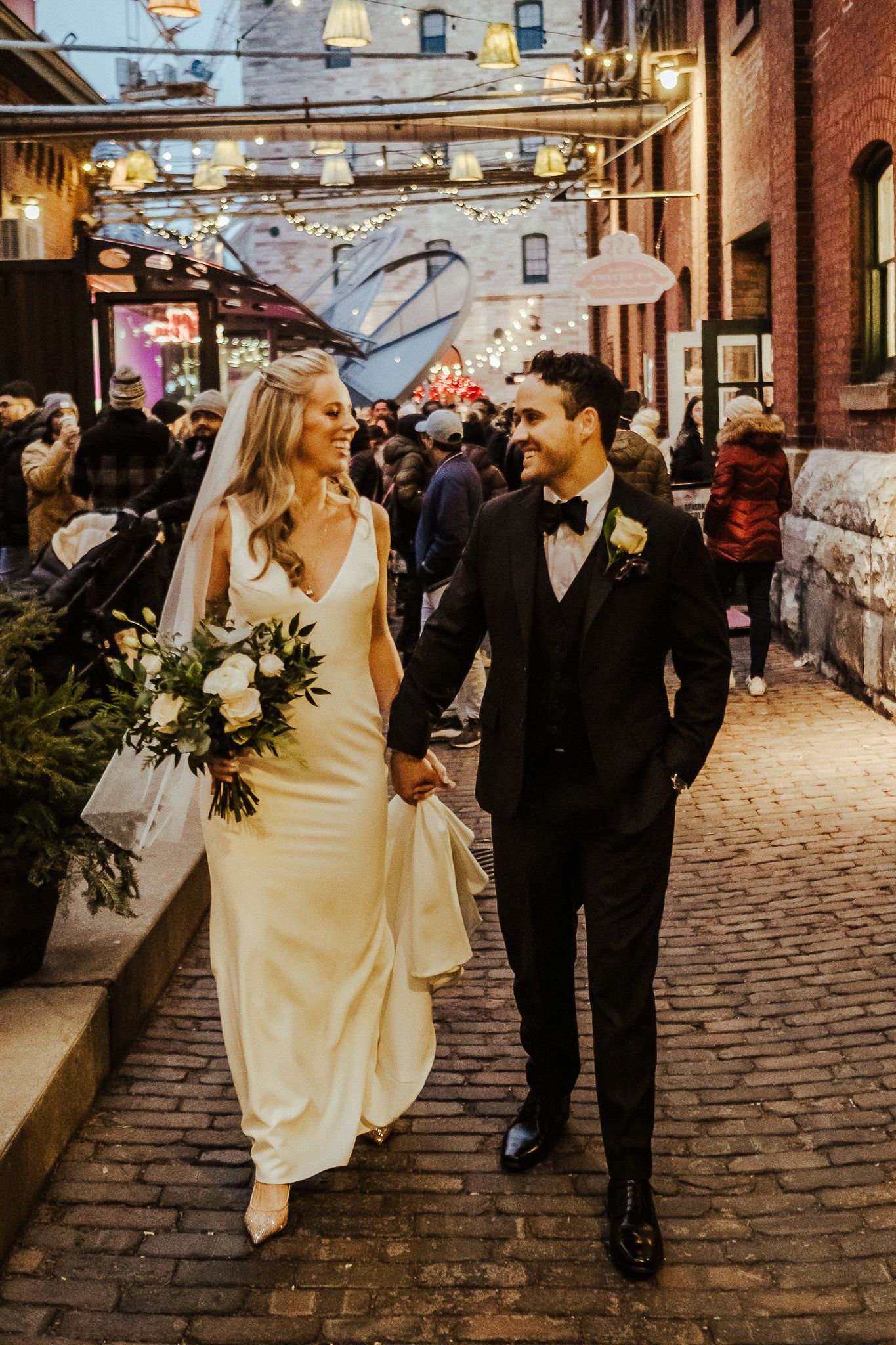 Couple walks down cobblestone street during golden hour with string lights overhead.