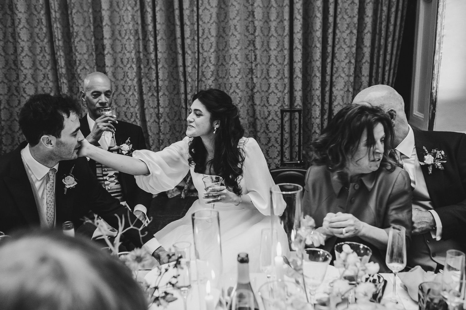 Candid black and white photo of wedding guests laughing and enjoying themselves at a reception dinner table.