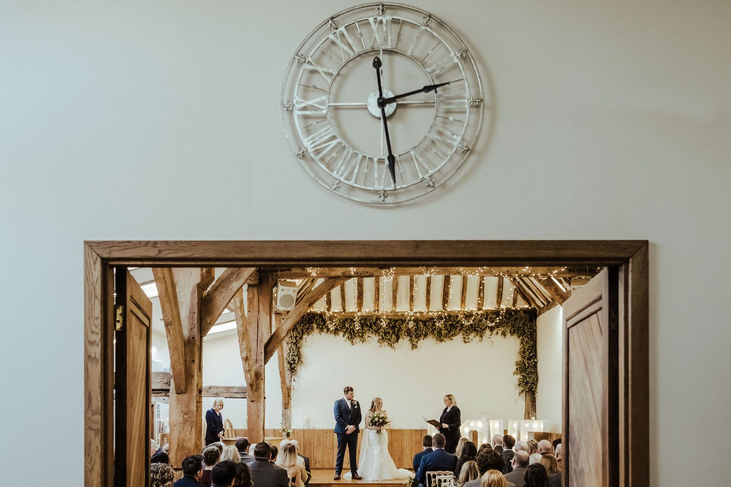 Wedding ceremony viewed through wooden doorframe with vintage clock above.