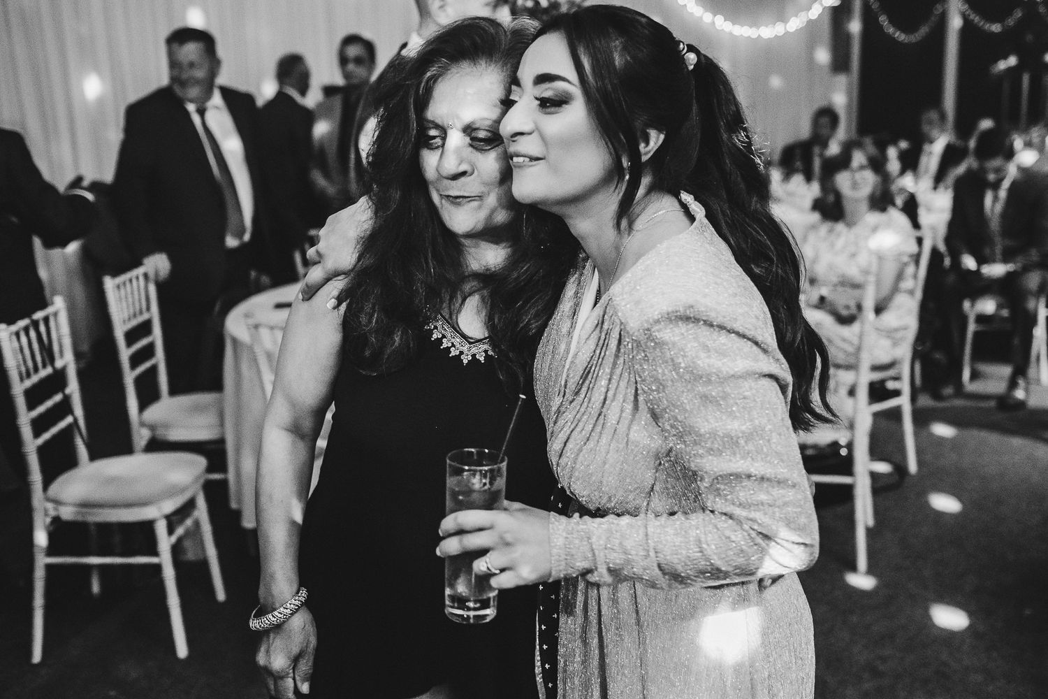 Two wedding guests in black and silver attire share an intimate moment at a reception with chairs in the background.
