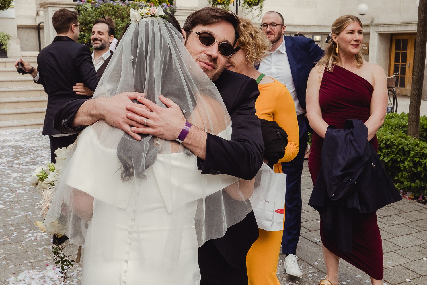 Wedding guests gather on stone steps for outdoor celebration as couple embraces during post-ceremony reception.