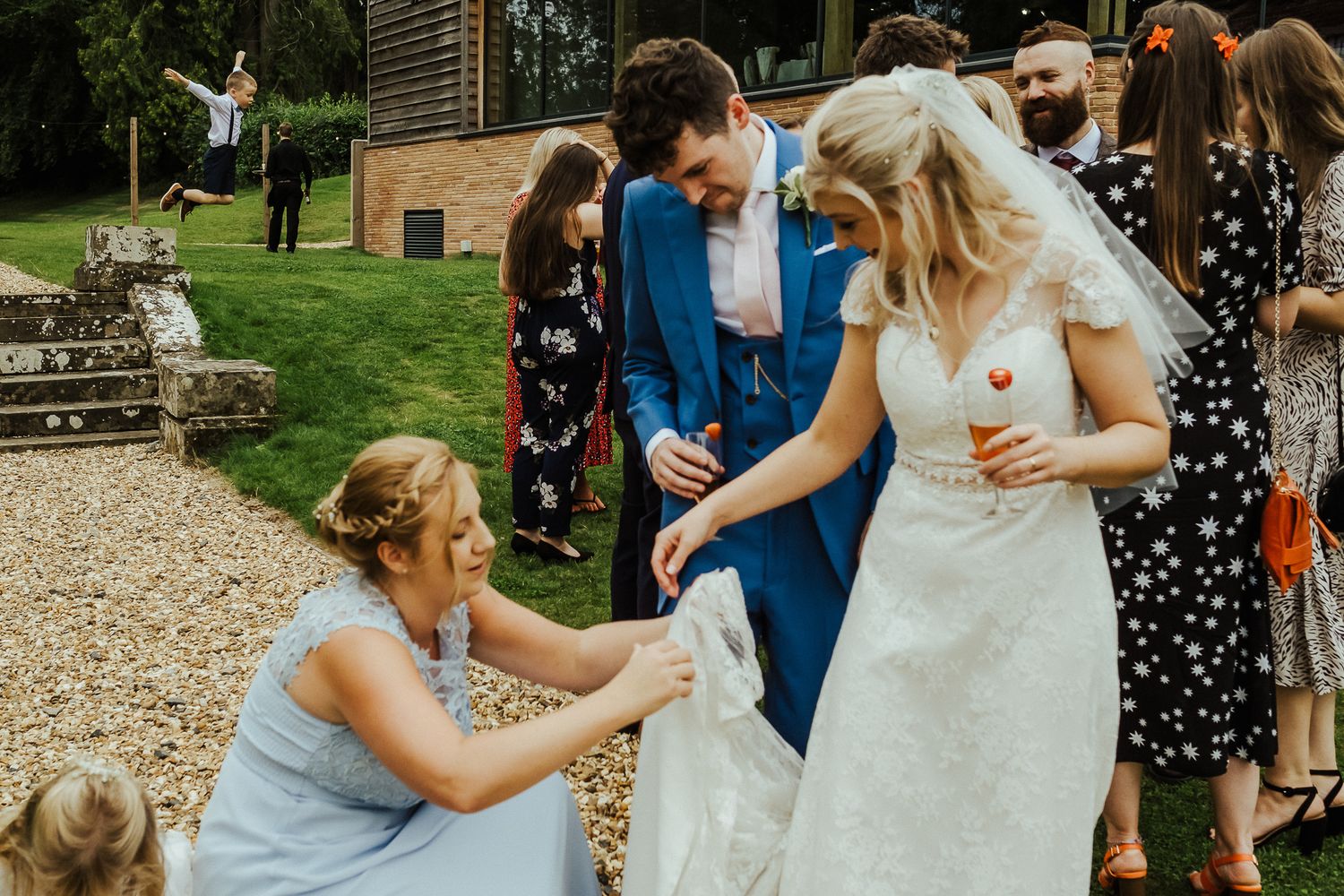 Wedding guests help adjust bride's dress train during outdoor garden reception.