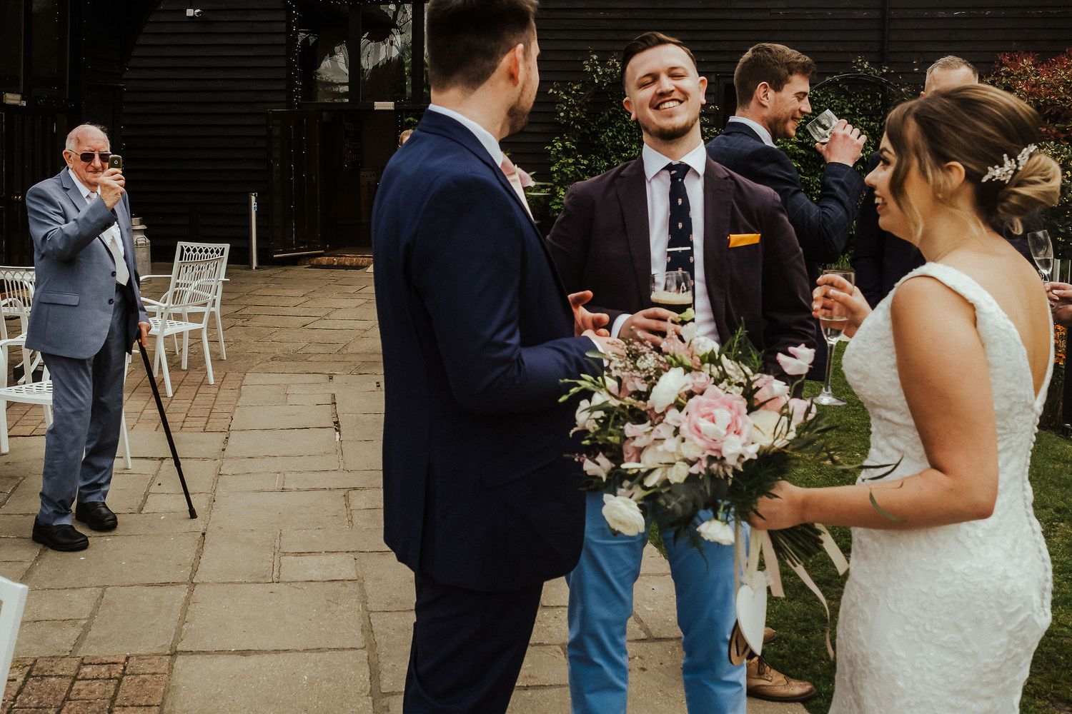 Wedding guests mingle and chat while holding bouquets in outdoor courtyard.
