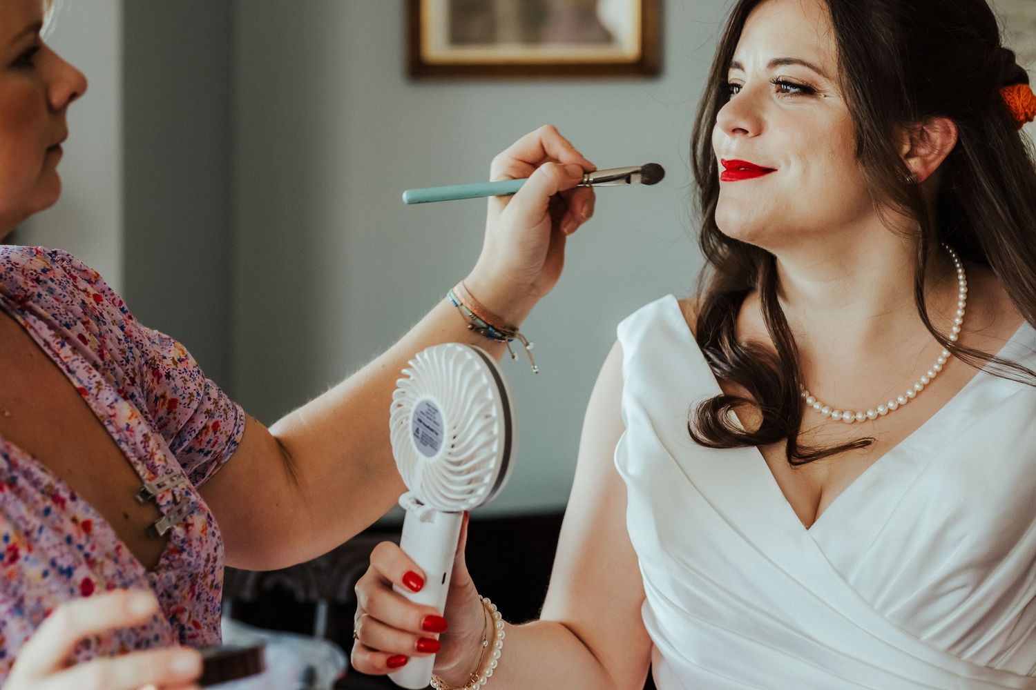 Makeup artist applies finishing touches to bridal look while holding a small fan.