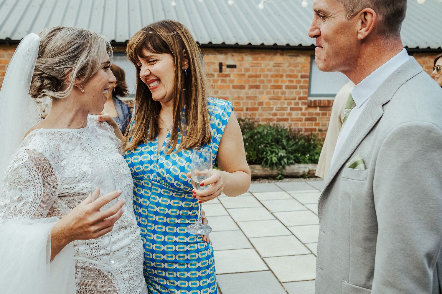 Wedding guests share joyful conversation during outdoor reception by brick building.