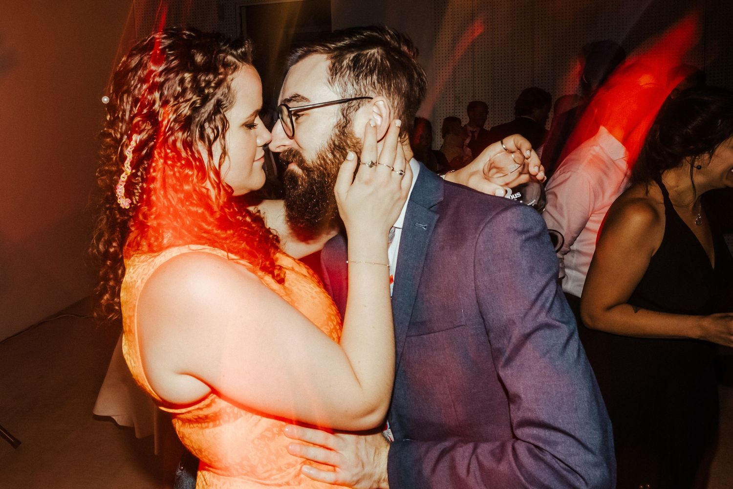 A couple shares an intimate dance moment illuminated by warm red lighting at a wedding reception.