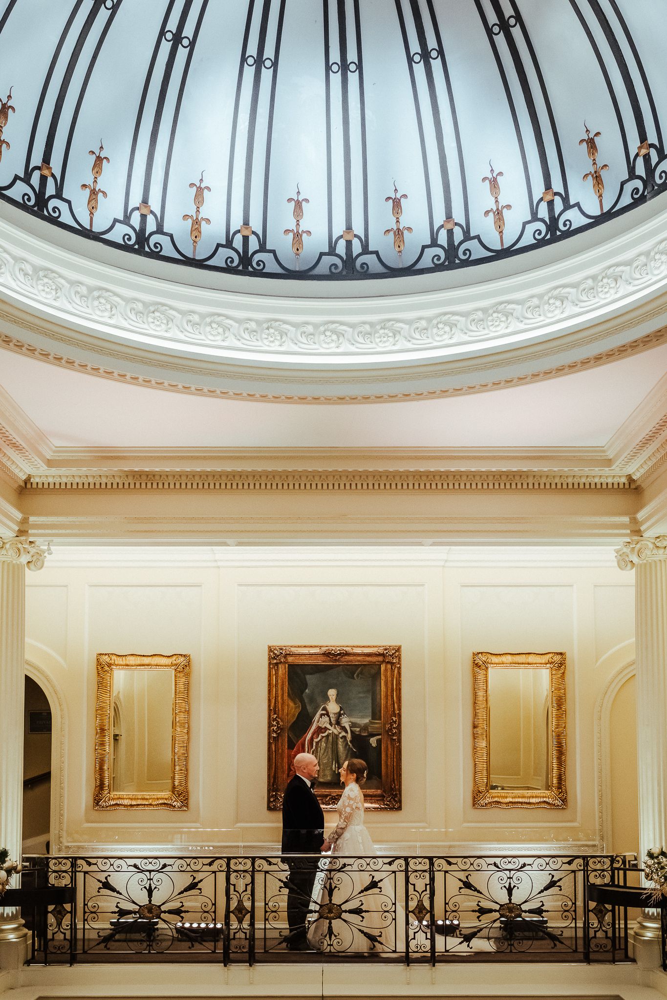 Couple stands beneath ornate domed ceiling with cross details in elegant interior setting.
